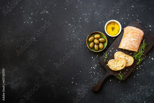Ciabatta bread on wooden board with olive oil, olives and herbs on black. Top view with copy space.