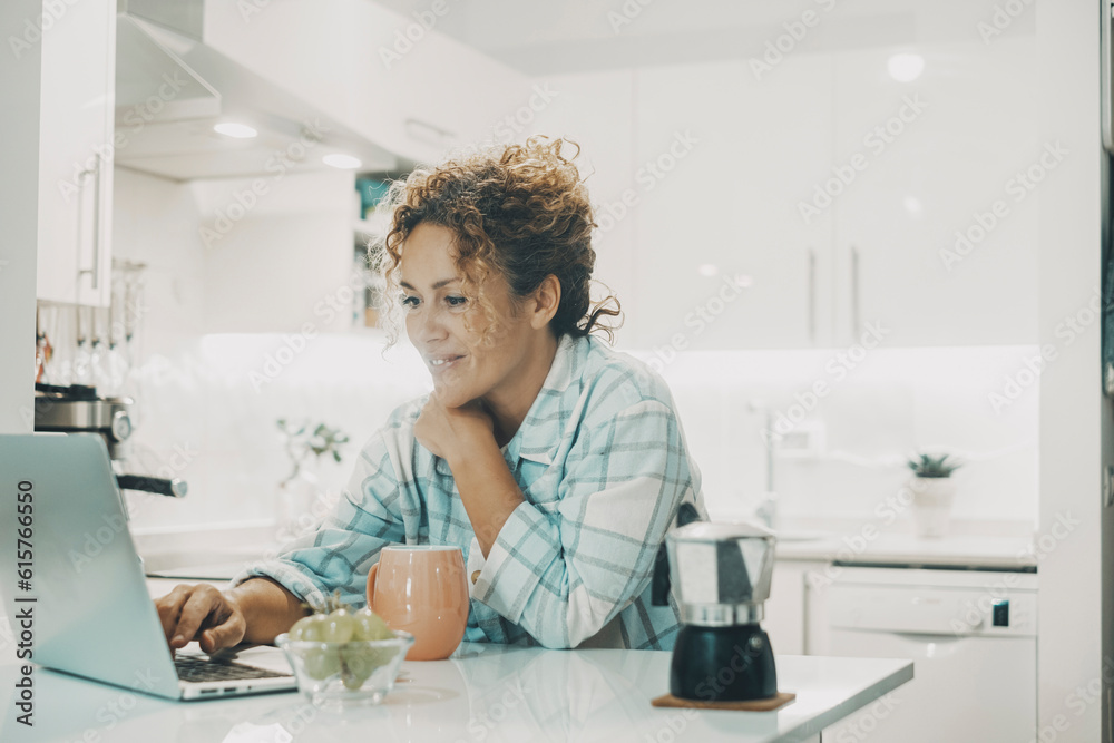 One modern young adult happy woman using laptop at home in the kitchen during morning breakfast and coffee time. Smart female people woriking on computer sitting inside apartment. Concept connection One modern young adult happy woman using laptop at home in the kitchen during morning breakfast and coffee time. Smart female people woriking on computer sitting inside apartment. Concept connection