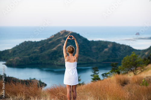 A young girl of European appearance, in a white dress, stands at the top with her hands folded in the shape of a heart above her head, and enjoys a beautiful view of the island and the bay. © Evgenii