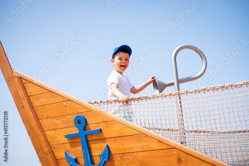 A beautiful cheerful happy child boy in a white t-shirt on an eco-friendly wooden playground in the form of a ship stands near the speaking tube photo