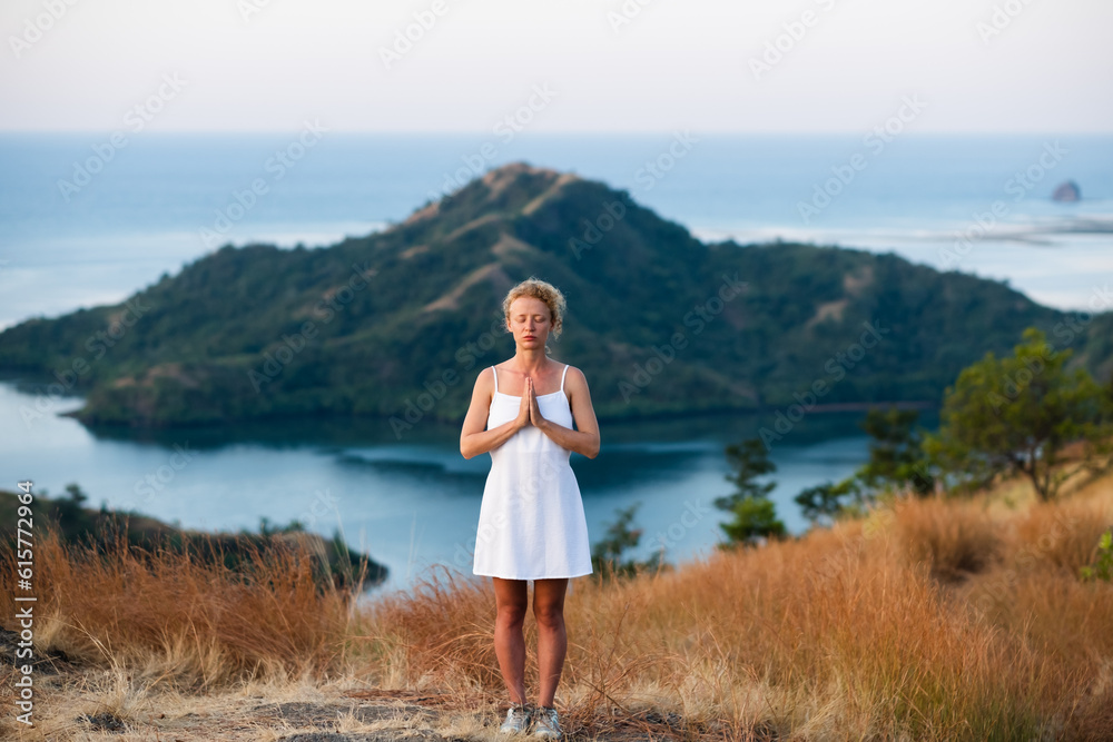 A beautiful, young girl of European appearance, with blond, curly hair, in a white dress, meditates with her hands clasped in namaste, against the backdrop of a beautiful landscape of islands and bays