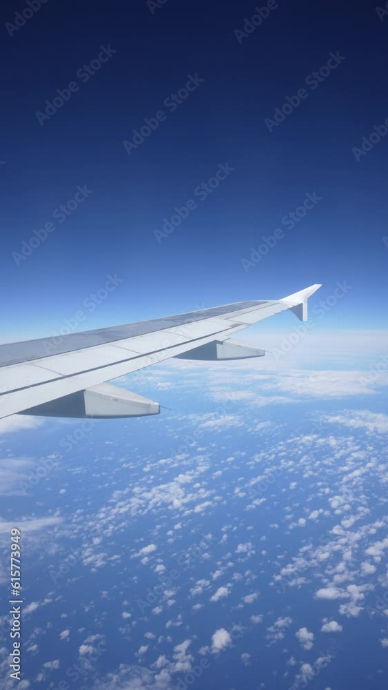 Vertical view through window on wing of plane flying above white bright clouds on background of blue sky on sunny day, slow motion. Slowly moving fluffy clouds from airplane window during flight. Vertical view through window on wing of plane flying above white bright clouds on background of blue sky on sunny day, slow motion. Slowly moving fluffy clouds from airplane window during flight.