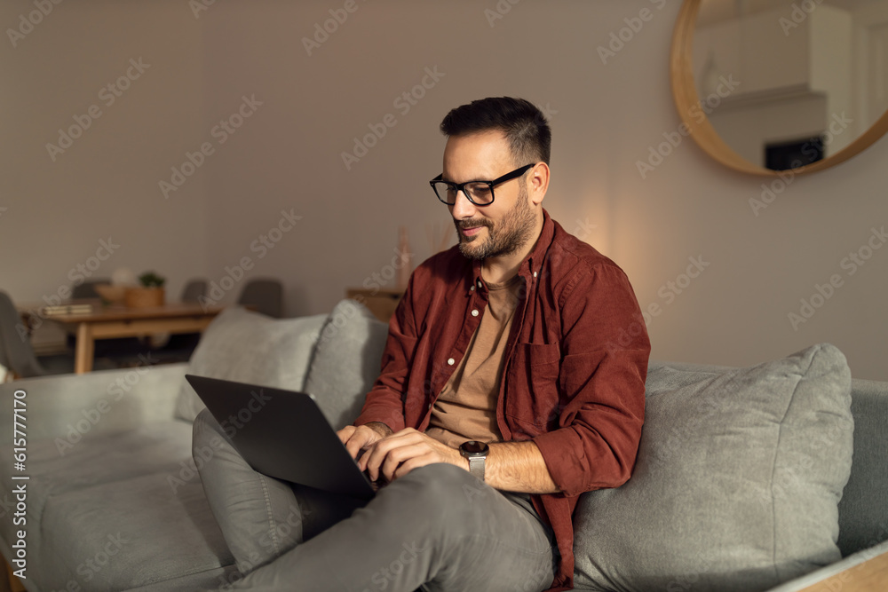 Relaxed man using a laptop, typing something, sitting on the sofa at home.