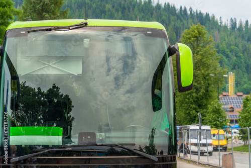 Close-up of a large windshield of a tourist bus at a bus stop, blurred background of green trees and buses, reflections of clouds and trees in the glass of the bus, summer travel