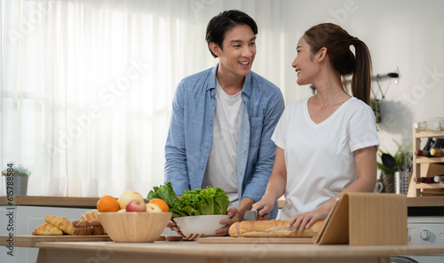Happy romantic couple together parepare breakfast in kitchen at home. Young Asian woman smiling cooking healthy meal of fresh fruit & vegetable for her family. Husband and wife morning diet lifestyle photo