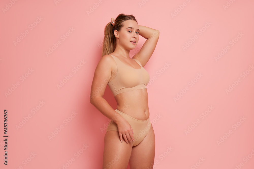 Body half-turned view of girl in sports bra and panties with one hand on her head another hand on her hip, poses on pink studio background, body comfort concept, copy space. - obrazy, fototapety, plakaty 