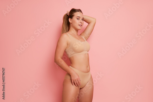 Body half-turned view of girl in sports bra and panties with one hand on her head another hand on her hip, poses on pink studio background, body comfort concept, copy space.