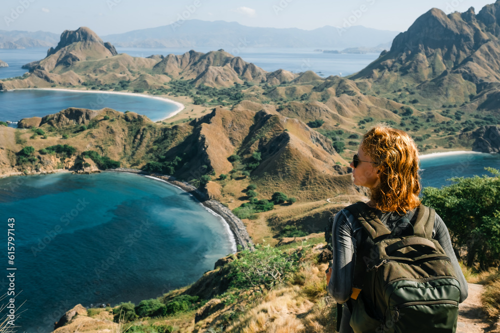 A young girl with a backpack stands on top of a mountain and admires the panorama of Padar Island in Indonesia, rear view.