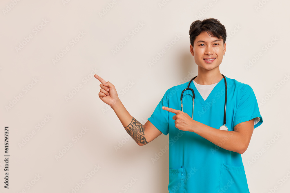 Young oriental male doctor in medical uniform poses with stethoscope on his neck against white backdrop and pointing fingers aside, professional people concept, copy space Young oriental male doctor in medical uniform poses with stethoscope on his neck against white backdrop and pointing fingers aside, professional people concept, copy space