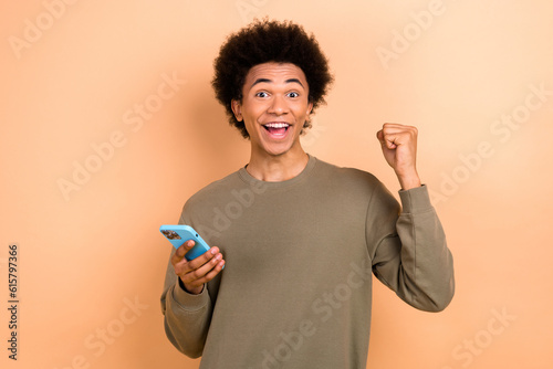 Photo of handsome delighted man hold smart phone raise fist celebrate success isolated on beige color background