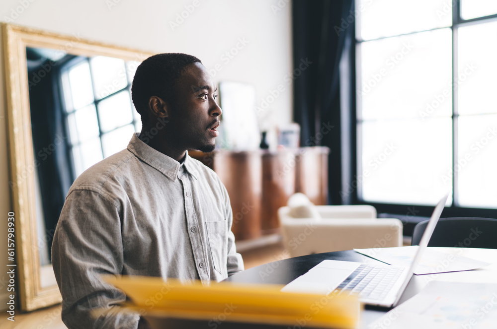 Serious black man sitting with laptop at table at home