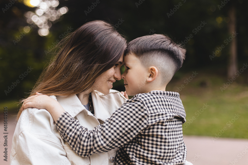Happy mom holding son boy in arms, hugging kid with noses touch, smiling, enjoying warm moment. Younger mum carrying kid and dancing in the park. Motherhood, adoption, family concept.
