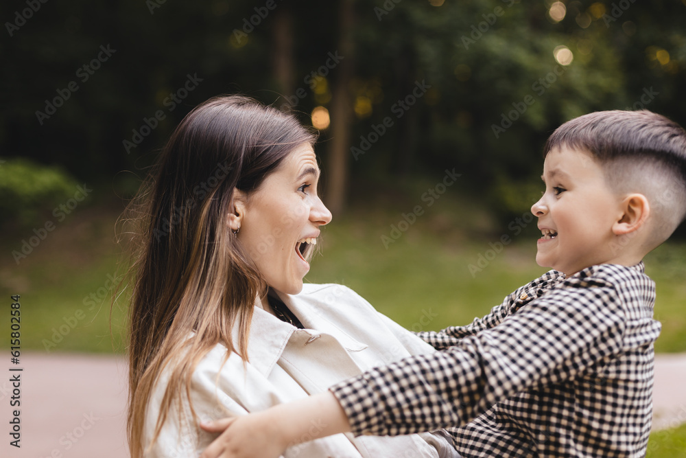 Happy mother twirling her son holding him on hands. Brunette mom with her son boy have a good time in park, Family day. Woman wear white jeans jacket, boy wear shirt.