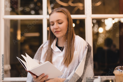 Photo of happy dreamy joyful young woman reading book, look dreaming and thoughtful sitting in cafe or restaurant. Dreaming blonde woman rest with book. Woman's shoulders cover by plaid. Focus on book