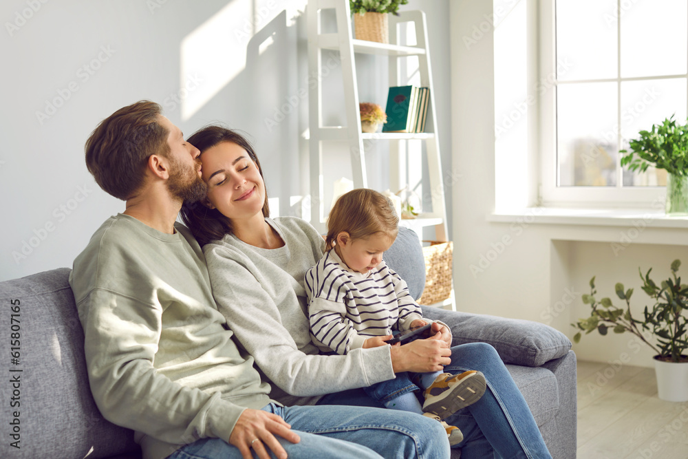Happy young family with little child girl with smartphone sitting on the sofa. Husband kissing his wife with tenderness while their daughter watching funny cartoon online on mobile phone.