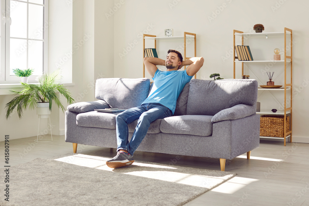 Young relaxed man sitting on comfortable couch in the living room at modern home and having a rest from a work with closed eyes. Calm peaceful guy enjoying quiet time alone in the room and dreaming.