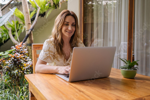 Young beautiful woman sitting in a garden with laptop in a tropical resort. Girl with laptop computer connecting to 4G wireless for browsing social media working remotely at terrace