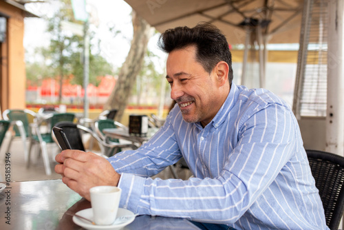 Happy mature latin man using cell phone sitting at street cafe