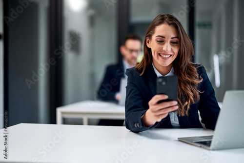A smiling female bank employee using a mobile phone while working over a laptop.