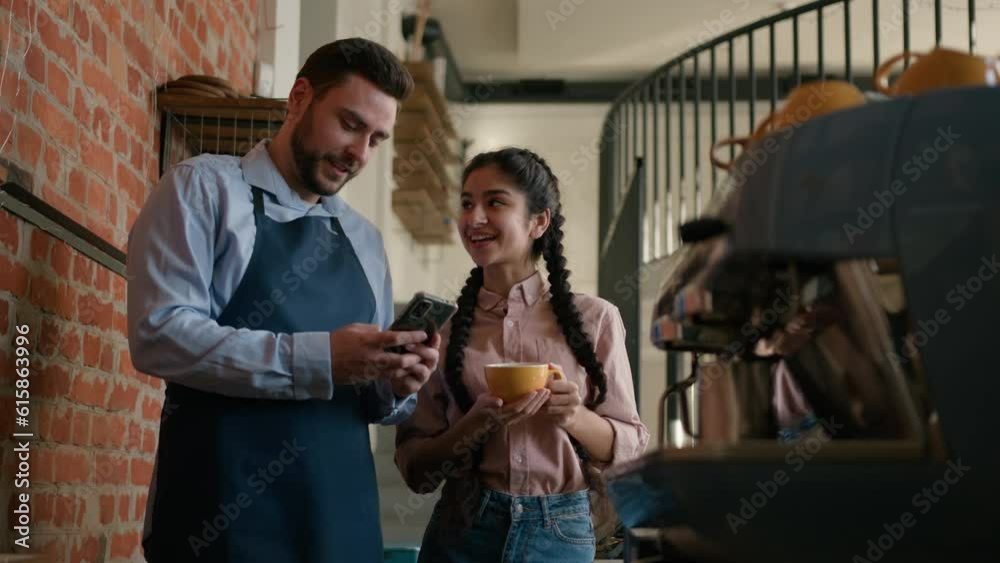 Caucasian man waiter bartender speaking to arabian woman restaurant manager behind counter talk on break in cafeteria browsing social media on phone colleagues work together communicate in free time