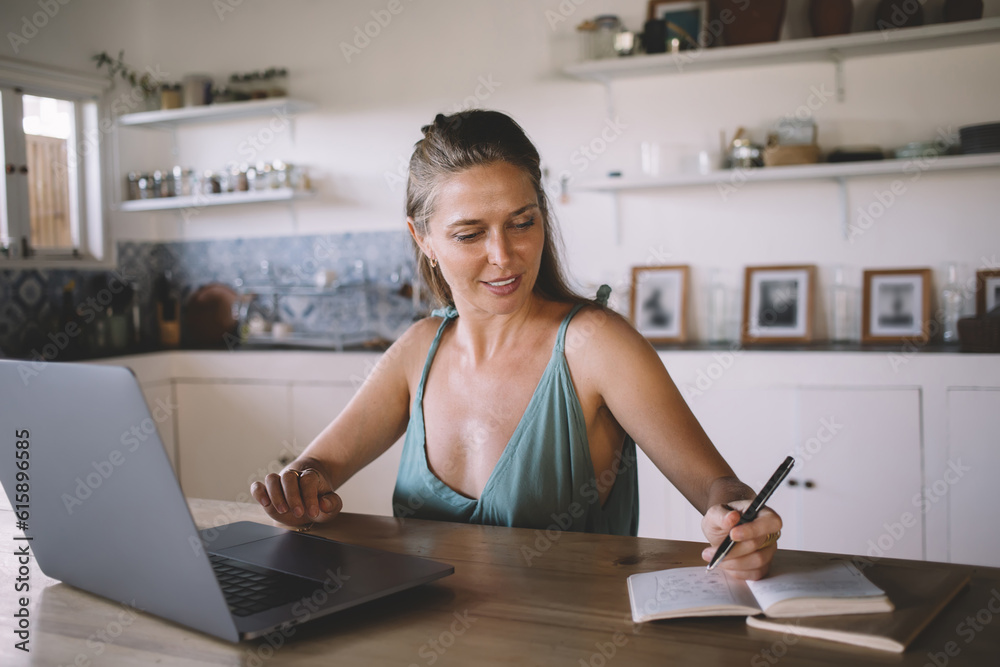 Smiling woman taking notes from information on laptop at table Smiling woman taking notes from information on laptop at table