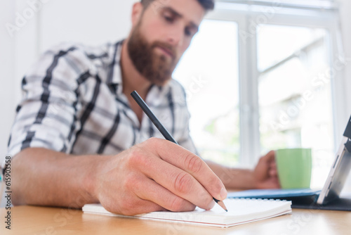Close-up of the hand of a creative young man holding a pencil over a blank notebook, while thinking of a new project or assignment during freelance work in a co-working space