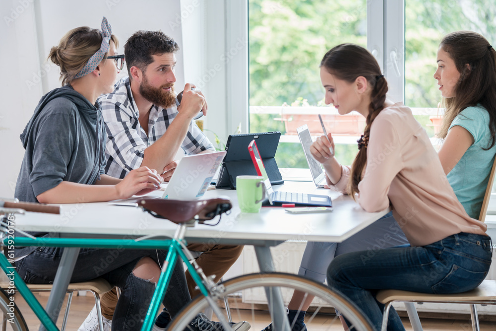 Four proficient freelancers and independent contractors co-working on tablets and laptops, connected to internet at a shared desk behind a commuter bike in a modern work hub