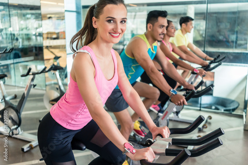 Side view of a beautiful fit young woman smiling while pedaling during cardio workout at indoor cycling group class in a modern fitness club