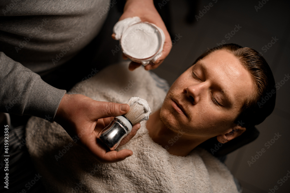 Fototapeta premium Barber applying shaving foam onto client's face using old-fashioned shaving brush in barbershop
