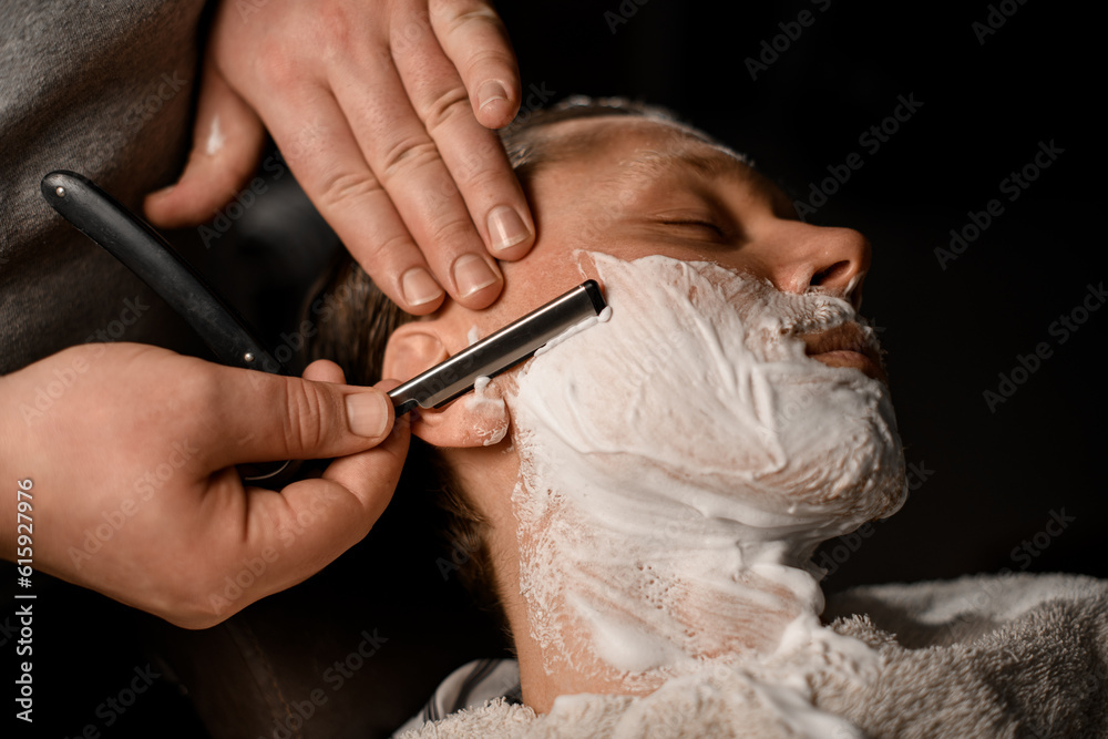 Barber man shaving young attractive client with vintage straight razor in modern barbershop, closeup view