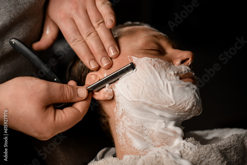 Barber man shaving young attractive client with vintage straight razor in modern barbershop  closeup view