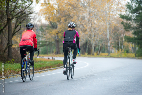 Two Young Female Cyclists Riding Road Bicycles in the Park in the Cold Autumn Morning. Healthy Lifestyle Concept.