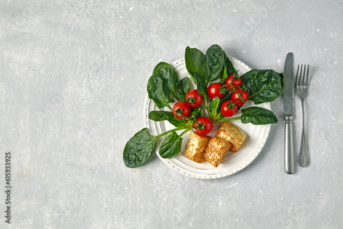Red cherry tomatoes on a branch, green spinach leaves and fried mozzarella curd cheese on a dark plate on a light concrete table. Top view with copy space