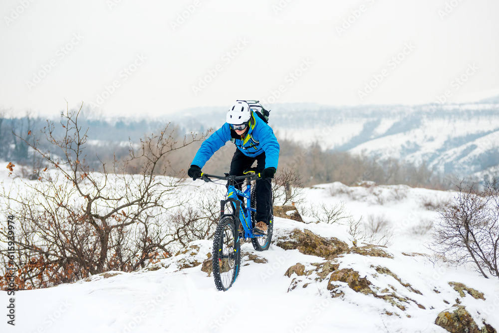 Cyclist in Blue Riding the Mountain Bike on the Rocky Winter Hill Covered with Snow. Extreme Sport and Enduro Biking Concept.