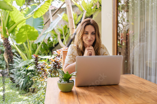 Young woman freelancer working on laptop computer sitting at table on wooden terrace of bungalow surrounded with greenery