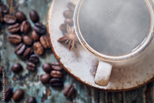 Hot steaming coffee in the cup on the old scratched table with coffee beans lying on it. View from above