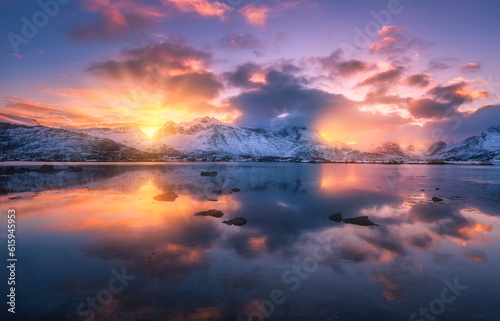 Sea coast  beautiful snowy mountains and colorful sky with clouds and golden sunlight at sunset in winter. Lofoten islands  Norway. Landscape  rocks in snow  reflection in water at dusk. Scenery