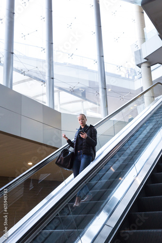 Young businesswoman with large black bag and mobile phone getting down on the escalator during business trip in modern city.