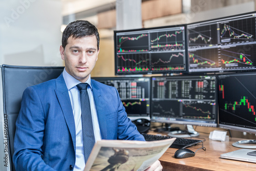 Business portrait of confident stocks broker holding a business newspaper in traiding office with multiple computer screens full of index charts and data analyses. photo