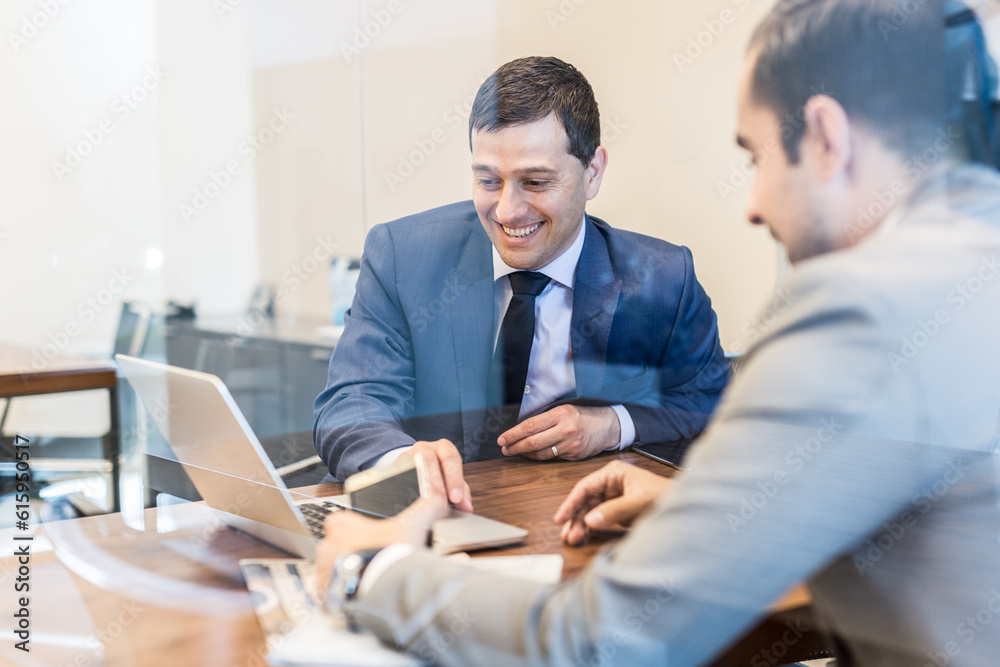 Through the window view of two cheerful young businessmen using laptop computer at business meeting in corporative office. Through the window view of two cheerful young businessmen using laptop computer at business meeting in corporative office.