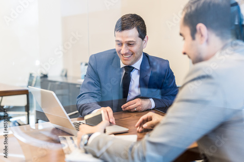 Φωτογραφία Through the window view of two cheerful young businessmen using laptop computer at business meeting in corporative office