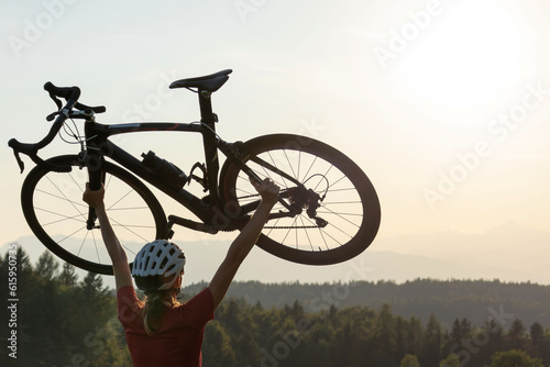 Female road cyclist raising bicycle with both arms above her head at sunset with beautiful green nature. Winning, success and achievement concept.