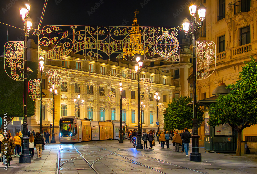 Fototapeta premium The Metrocentro system Tram travels through the historic downtown at night under holiday decorations with the Gothic Seville Cathedral in view in Seville, Spain.