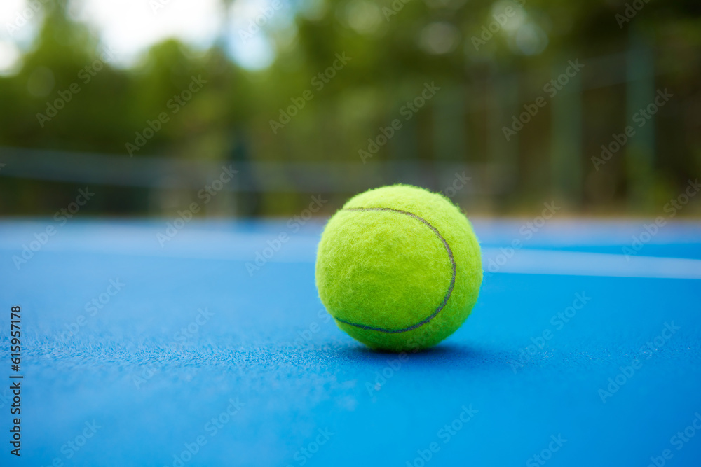 Yellow ball is laying on blue tennis court background. Blurred green plantings and trees behind. Contrast image with satureted colors. Concept of sport equipment photo.