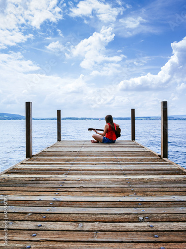 Photo of a young woman playing her acoustic guitar on a dock on a lake.