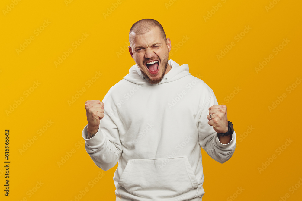 Bearded man wearing white hoodie raising fists up acts like he won something. Lucky young hipster man isolated over yellow background. Bearded man wearing white hoodie raising fists up acts like he won something. Lucky young hipster man isolated over yellow background.