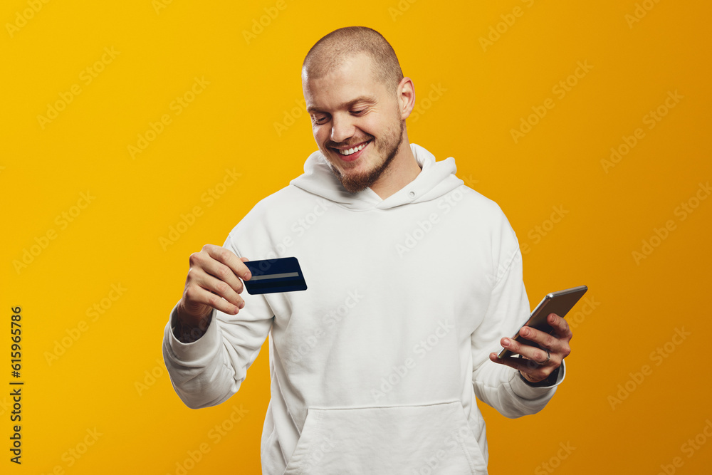 Smiling bearded man wears white hoodie using mobile cell phone hold credit bank card doing online shopping order delivery isolated over yellow wall Smiling bearded man wears white hoodie using mobile cell phone hold credit bank card doing online shopping order delivery isolated over yellow wall