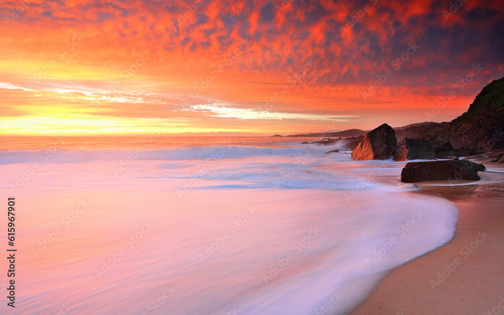 Obraz premium Foamy white seas wash onshore and above the skies turn on brilliant hues of yellow to red