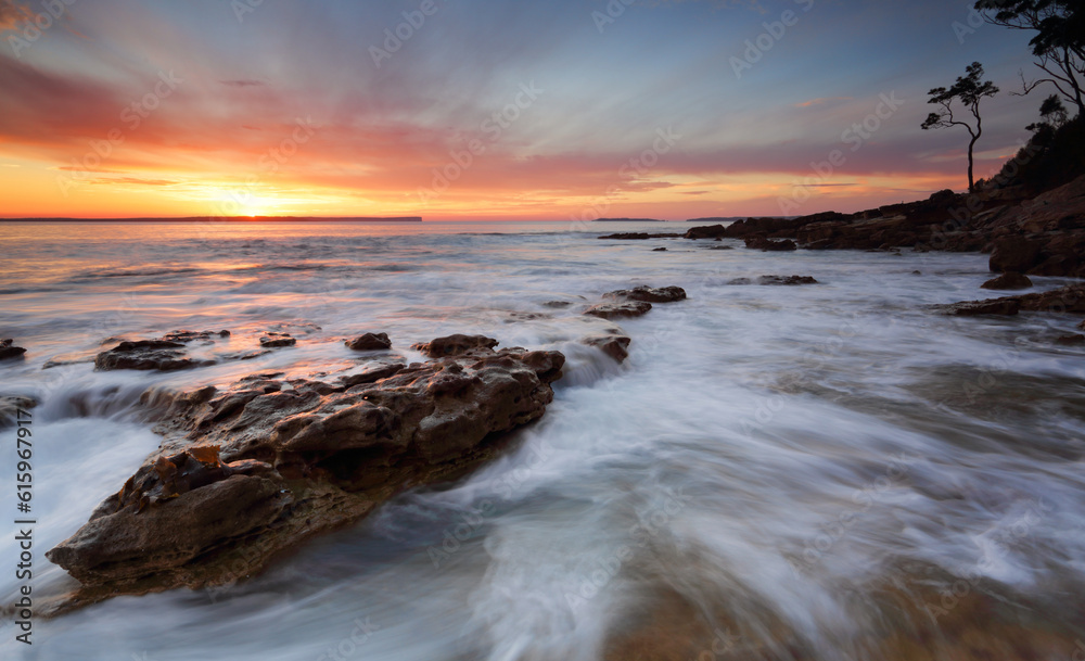 Beautifuol sunrise over the bay as the ocean ebbs and flows gently over rocks at the shore edge