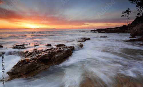 Beautifuol sunrise over the bay as the ocean ebbs and flows gently over rocks at the shore edge